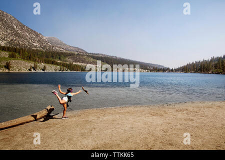 Ragazza in piedi su una gamba sola a riverbank contro le montagne e il cielo chiaro Foto Stock