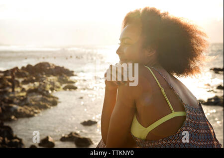 Riflessivo sorridente ragazza seduta sulla riva del mare durante la giornata di sole Foto Stock