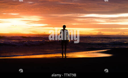 Lunghezza completa di silhouette turistico con tavola da surf guardando il mare mentre in piedi sulla riva contro il cielo durante il tramonto Foto Stock