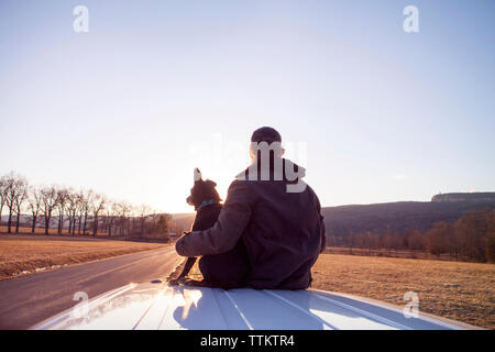 Vista posteriore dell'uomo seduto con il cane sul tetto del veicolo contro il cielo chiaro Foto Stock