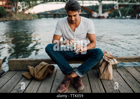 Apertura uomo può bere stando seduti sul molo sul fiume durante il tramonto Foto Stock