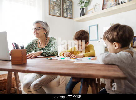 Ragazzi facendo disegni mentre la nonna utilizzando portatile a casa Foto Stock