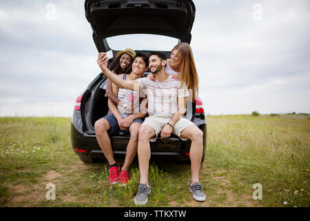 Happy amici prendendo selfie attraverso smart phone mentre sedendo nel baule auto sul campo contro sky Foto Stock