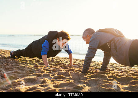 Determinato il padre e il figlio facendo push-up insieme contro il cielo chiaro in spiaggia durante il tramonto Foto Stock