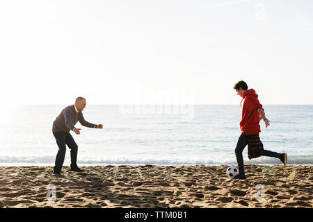 Figlio di calci palla calcio mentre padre difendere presso la spiaggia contro il cielo chiaro Foto Stock