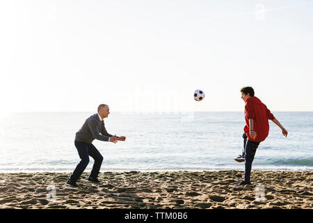 Giocoso figlio di calci palla calcio mentre padre difendere presso la spiaggia contro il cielo chiaro Foto Stock