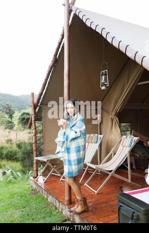 Donna avvolta in asciugamano di bere il caffè mentre si sta in piedi in tenda durante le vacanze Foto Stock