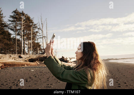 Vista laterale della donna fotografando attraverso smart phone sulla spiaggia Foto Stock