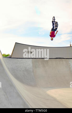 Maschio con rider BMX bike eseguendo backflip sulla rampa in calcestruzzo contro il cielo nuvoloso a skate park Foto Stock