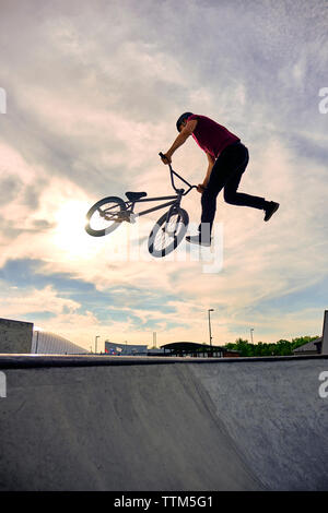 Basso angolo di visione del pilota maschio con biciclette BMX eseguendo tailwhip sulla rampa in calcestruzzo contro il cielo nuvoloso Foto Stock