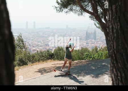 Vista posteriore della donna a braccia alzate con lo skateboard su strada contro sky Foto Stock