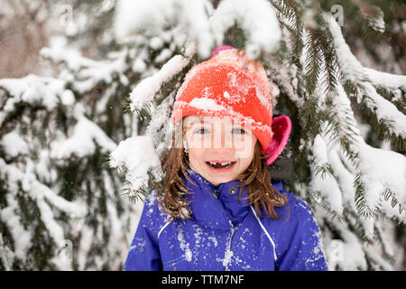 Ritratto di Allegro ragazza di indossare un abbigliamento caldo mentre in piedi contro la coperta di neve tree Foto Stock