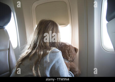 Vista posteriore della ragazza con Teddy bear guardando attraverso la finestra mentre si viaggia in aereo Foto Stock