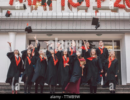 Basso angolo vista di allievi felici di indossare abiti di graduazione permanente sulla procedura da costruire Foto Stock