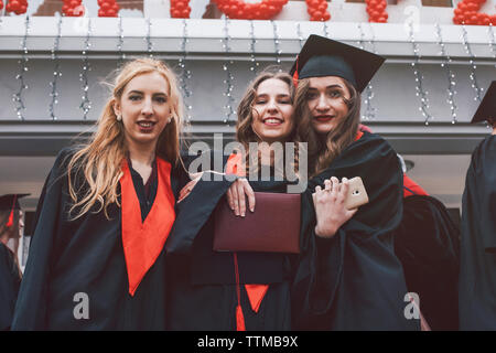 Angolo basso ritratto di felice studentesse indossando abiti di graduazione contro la costruzione di Foto Stock