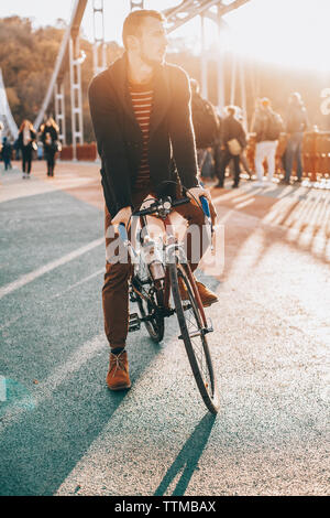 Ciclista maschio che guarda lontano mentre equitazione Bicicletta sul ponte in città durante il tramonto Foto Stock