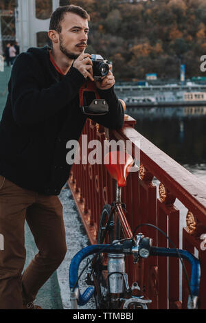 Ciclista a fotografare con la fotocamera mentre in piedi con la bicicletta sul ponte in città durante il tramonto Foto Stock