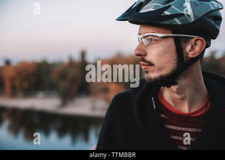 Close-up di pensosa ciclista indossando il casco e occhiali in piedi contro il cielo durante l'autunno Foto Stock