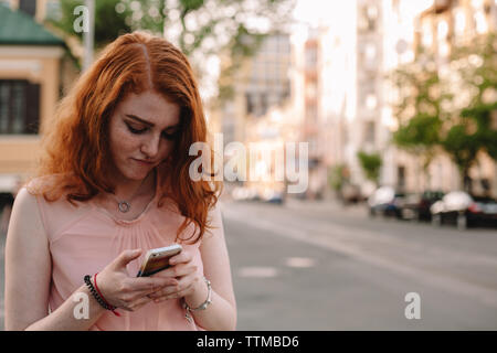 Carino adolescente utilizzando smart phone mentre in piedi in strada di città Foto Stock