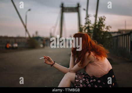 Giovane donna redheaded Sigaretta fumare mentre è seduto sul ponte Foto Stock