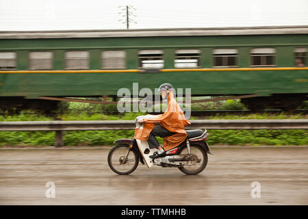 Il Vietnam, Hanoi, Campagna, un uomo passa un treno sul suo ciclomotore sotto la pioggia Foto Stock