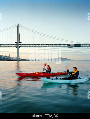 Stati Uniti, California, San Francisco, un uomo e una donna kayak nella Baia di San Francisco sotto il ponte della baia e la città di San Francisco in distanza Foto Stock