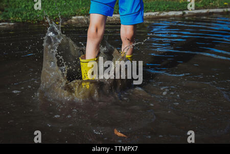 Sezione bassa di giocoso boy stamping piede mentre in piedi in una pozza in posizione di parcheggio Foto Stock