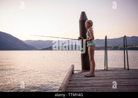 Vista laterale della ragazza la pesca nel lago mentre si sta in piedi sul molo contro il cielo chiaro durante il tramonto Foto Stock