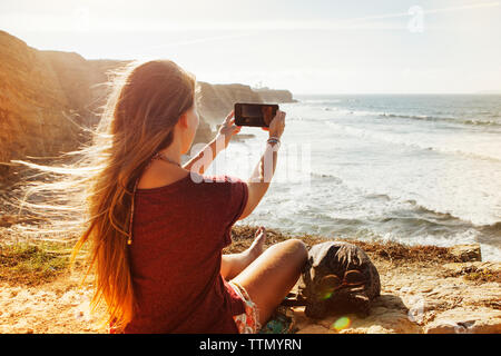 Vista posteriore della donna fotografare mentre è seduto sulla scogliera dal mare Foto Stock