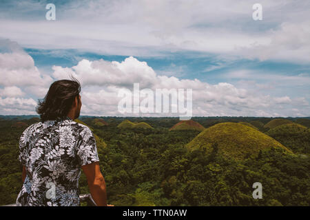 Vista posteriore di un uomo guarda il Chocolate Hills mentre in piedi dalla ringhiera contro il cielo nuvoloso Foto Stock