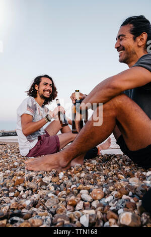 Allegro amici tenendo le bottiglie di alcol mentre è seduto sulla ghiaia a beach contro sky Foto Stock