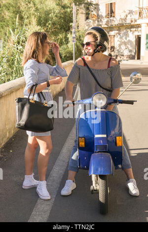 Ragazza adolescente parlando a femmina amico seduto su scooter in città Foto Stock