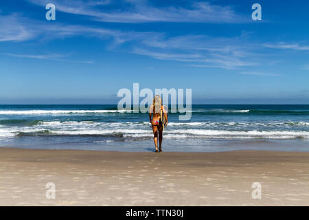Vista posteriore della donna in bikini che trasportano le tavole da surf mentre si cammina verso il mare contro il cielo blu durante la giornata di sole Foto Stock