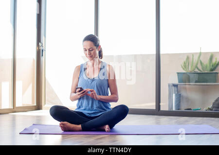 Donna che utilizza smart phone mentre è seduto sul tappeto di esercizio a casa Foto Stock