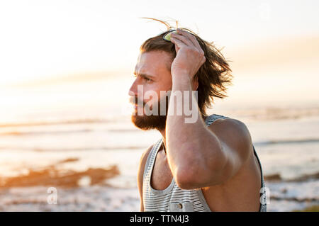 Vista laterale di pensosa uomo la regolazione degli occhiali da sole in spiaggia durante il tramonto Foto Stock