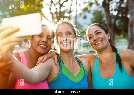 Happy amici di sesso femminile prendendo selfie con il cellulare mentre in piedi in park durante il tramonto Foto Stock