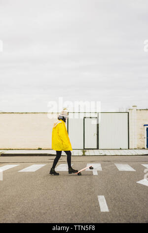 Vista laterale della giovane donna con lo skateboard indossando un impermeabile giallo sulla strada contro il cielo in città Foto Stock