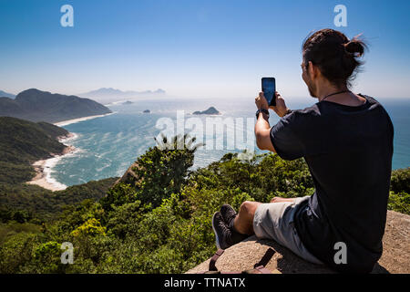 Lunghezza completa di fotografare turistico mare con smart phone mentre è seduto sulla scogliera contro sky durante la giornata di sole Foto Stock