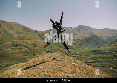 Vista posteriore della donna felice con le braccia sollevate il salto sulla montagna contro il cielo chiaro durante la giornata di sole Foto Stock