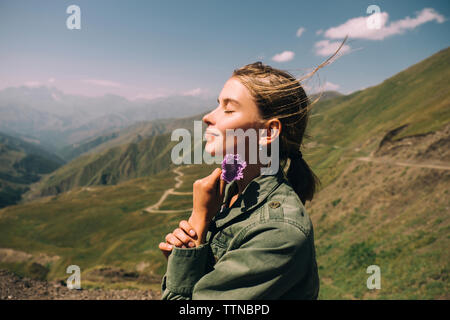 Vista laterale della donna sorridente con gli occhi chiusi fiore di contenimento mentre si sta in piedi sul monte contro sky durante la giornata di sole Foto Stock