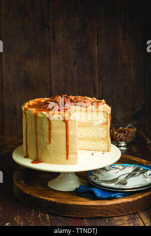 Torta al caramello su cakestand con piatti e forchette contro il muro di legno a casa Foto Stock