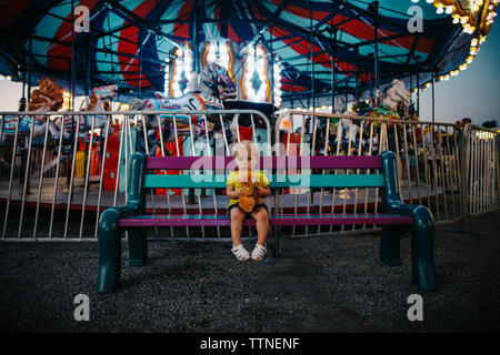 Ritratto di bambina giocattolo di contenimento mentre è seduto su un banco di lavoro presso il parco di divertimenti Foto Stock