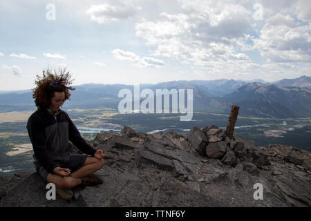 Lunghezza completa dell'uomo meditando mentre è seduto sulla roccia contro sky Foto Stock