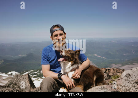 Ritratto di uomo con il cane seduto sul monte contro sky Foto Stock