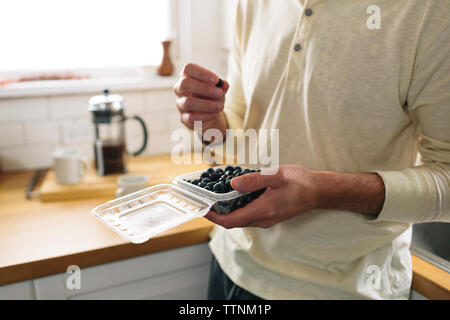 Sezione mediana dell'uomo della scatola di contenimento con mirtilli mentre appoggiata sul banco di cucina a casa Foto Stock