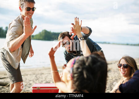 Happy amici facendo alta cinque mentre godendo a riverbank contro il cielo nuvoloso Foto Stock