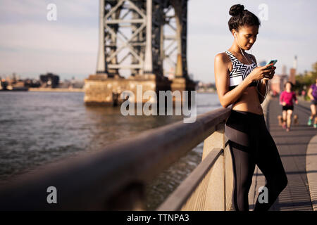 Donna che utilizza il telefono mentre si ascolta la musica dalla ringhiera sul lungomare Foto Stock