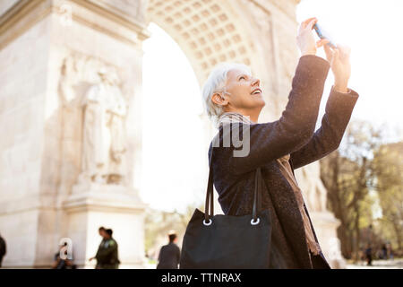 Vista laterale della donna senior fotografando attraverso smart phone mentre in piedi contro un arco trionfale in città Foto Stock