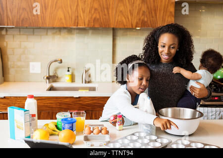 La madre che porta il figlio guardando la figlia rendendo i tortini in cucina Foto Stock