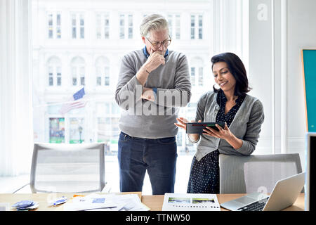 donna d'affari che mostra un computer tablet a un uomo d'affari anziano mentre si trova davanti a una finestra nell'ufficio creativo Foto Stock
